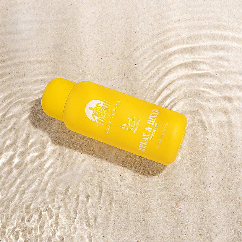 A bright yellow bottle of UV Block SPF 50+ Water-Based Sunscreen by The Beach Collective, placed diagonally on a textured sandy surface with ripples of shallow water.