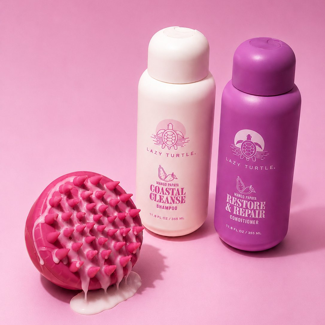 Three Lazy Turtle haircare products and a pink silicone scalp massager arranged on a pink background: a white Coastal Cleanse bottle, a purple Restore & Repair conditioner bottle, and a pink scrubber with white product dripping from it.