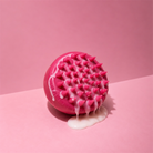 Pink silicone scalp brush with pointed bristles and white lather against a pink background.